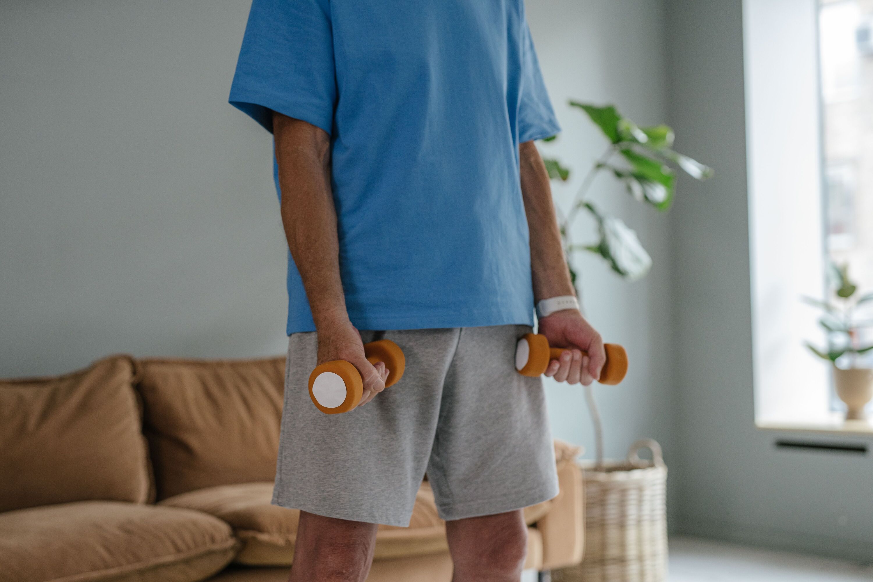 Man in a blue T-shirt holding two light orange dumbbells in a bright living room