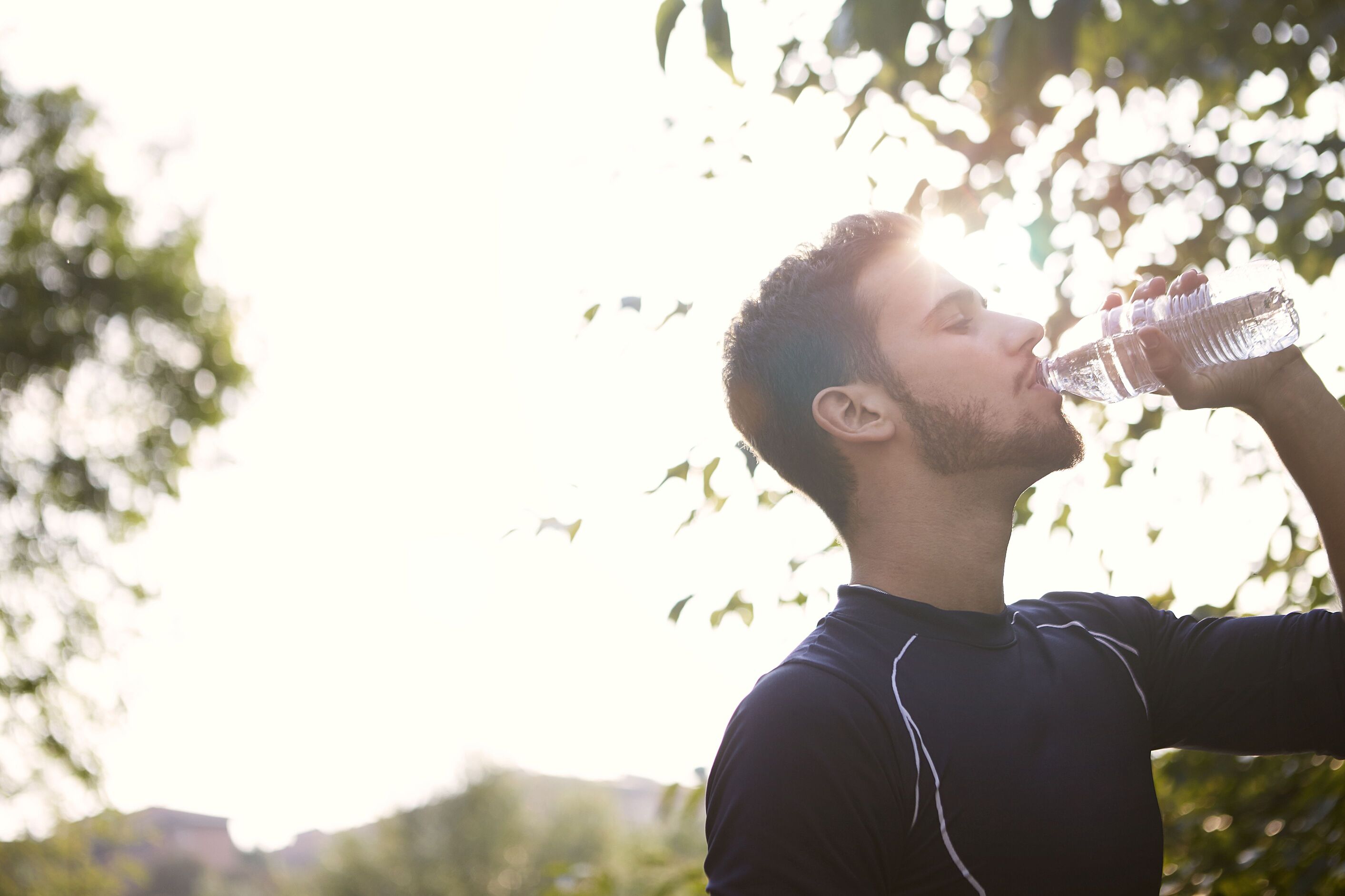 Man drinking water from a bottle outdoors under bright sunlight and trees